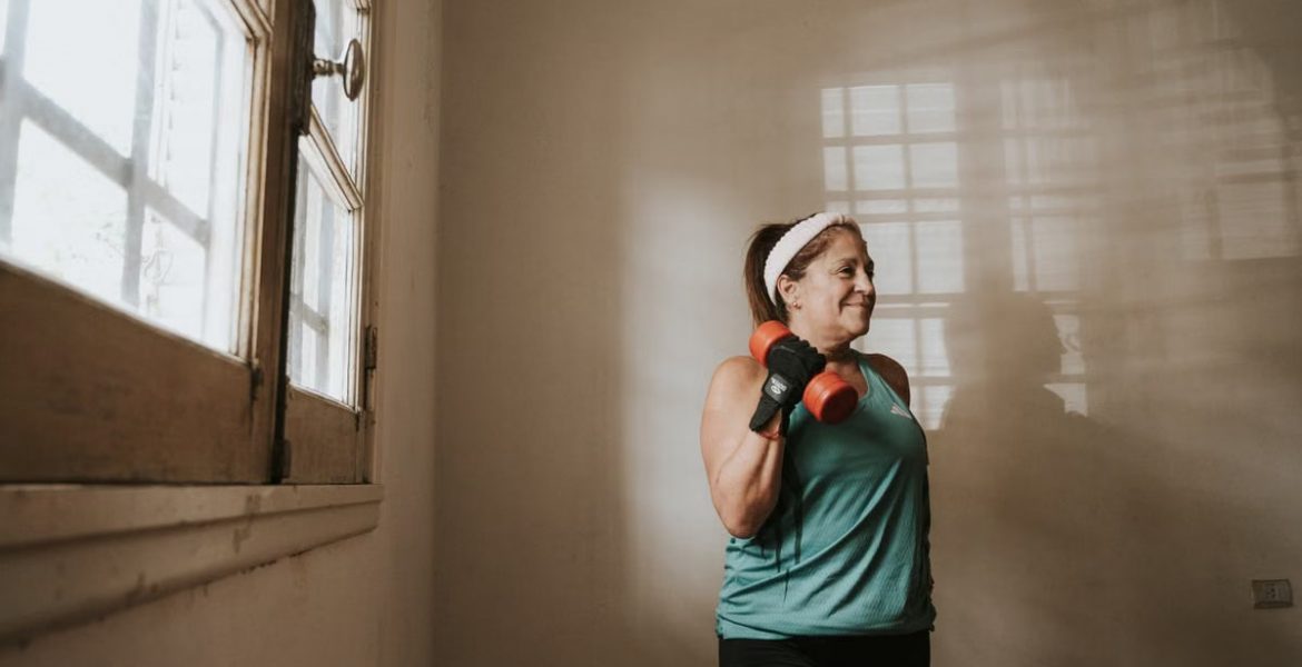Senior woman in a brightly lit room exercises with dumbbells, promoting fitness at home.