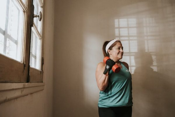 Senior woman in a brightly lit room exercises with dumbbells, promoting fitness at home.