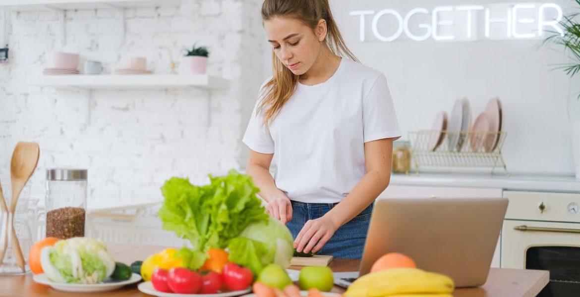 Woman in white t-shirt preparing fresh vegetables in a bright kitchen with a laptop.