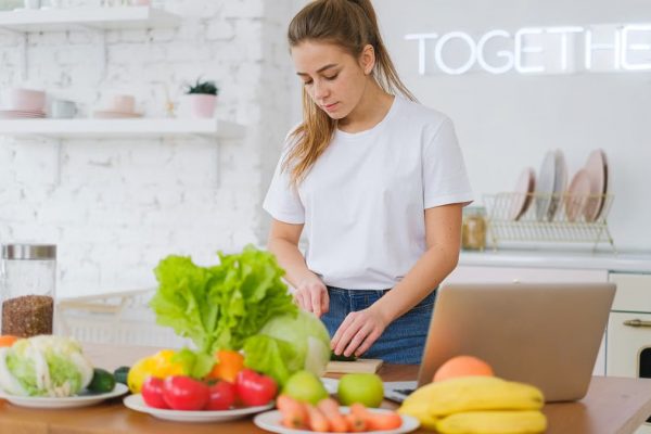 Woman in white t-shirt preparing fresh vegetables in a bright kitchen with a laptop.