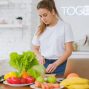 Woman in white t-shirt preparing fresh vegetables in a bright kitchen with a laptop.